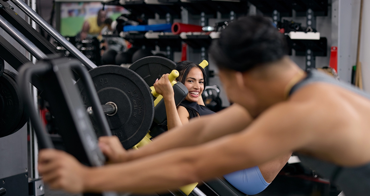 A woman smiling while using a leg press machine in a gym, with another person working out on a nearby machine in the foreground.